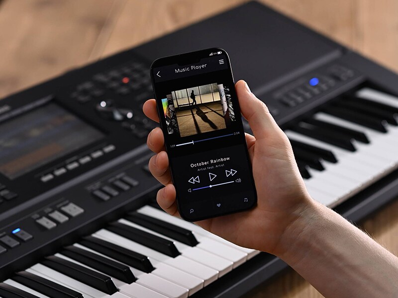Đàn organ Yamaha Arrange Workstation PSR-SX920 và một bàn tay đang cầm điện thoại thông minh có màn hình trình phát nhạc.
