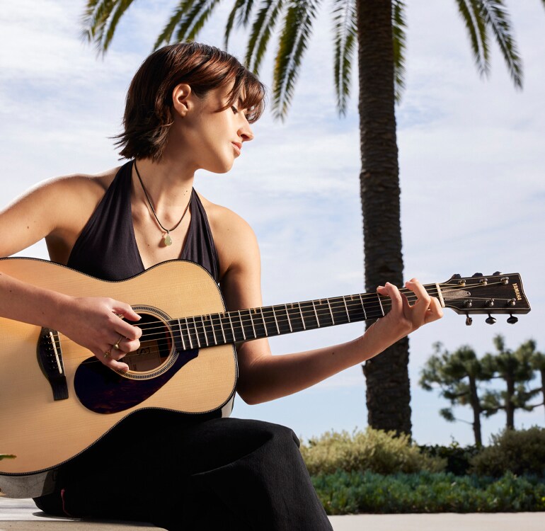 Guitarist Sophia James playing an FS9 guitar sits in front of a palm tree outside and plays an FS9 acoustic guitar.